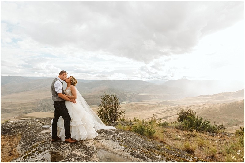 Snohomish & seattle wedding photographer | gsquared weddings photography a bride and groom embrace and kiss on a rocky ledge during their methow elopement overlooking a vast landscape of rolling hills beneath a cloudy sky She wears a white gown and hes in a vest and dress shirt | wedding photo by gsquared weddings