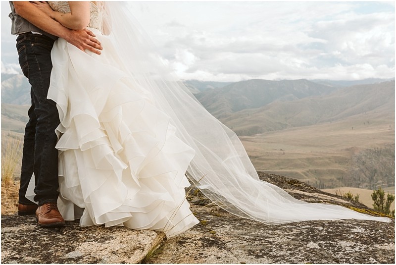 Snohomish & seattle wedding photographer | gsquared weddings photography a bride in a ruffled white gown and long veil embraces her groom on rocky ground surrounded by distant hills and cloudy skiesa romantic methow elopement set against a breathtaking mountain landscape Only their lower bodies are visible | wedding photo by gsquared weddings