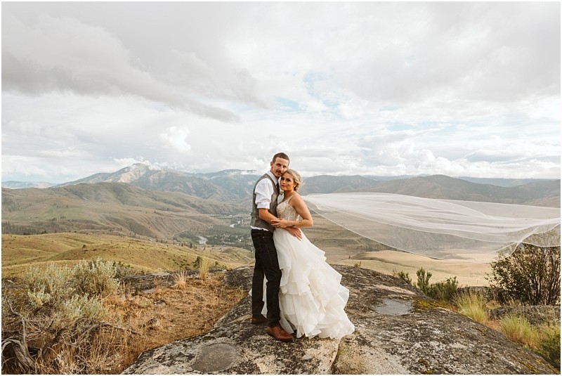Snohomish & seattle wedding photographer | gsquared weddings photography a bride and groom stand on a rocky hilltop with mountains in the background during their methow elopement The brides veil flows in the wind as they embrace surrounded by grassy hills and a cloudy sky | wedding photo by gsquared weddings