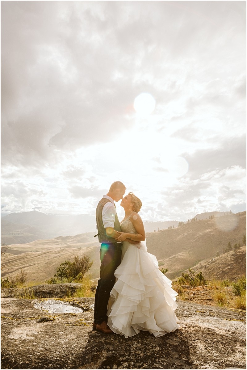Snohomish & seattle wedding photographer | gsquared weddings photography a bride and groom embrace on a rocky hilltop at sunset during their methow elopement with dramatic clouds and sunlight illuminating the landscape The bride wears a flowing white gown and the groom is in a vest and dress pants | wedding photo by gsquared weddings