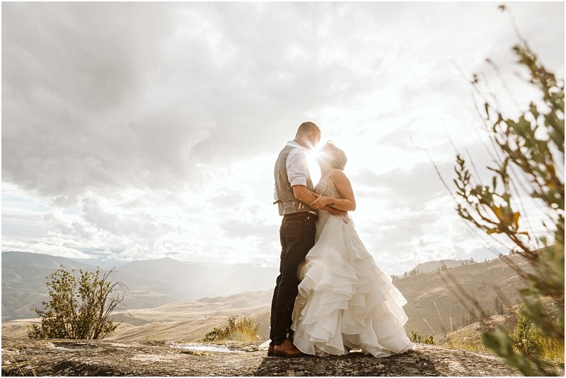 Snohomish & seattle wedding photographer | gsquared weddings photography a bride and groom embrace on a rocky hilltop at sunset during their methow elopement with sunlight streaming between them and mountains in the background The scene is romantic and scenic with a dramatic sky overhead | wedding photo by gsquared weddings