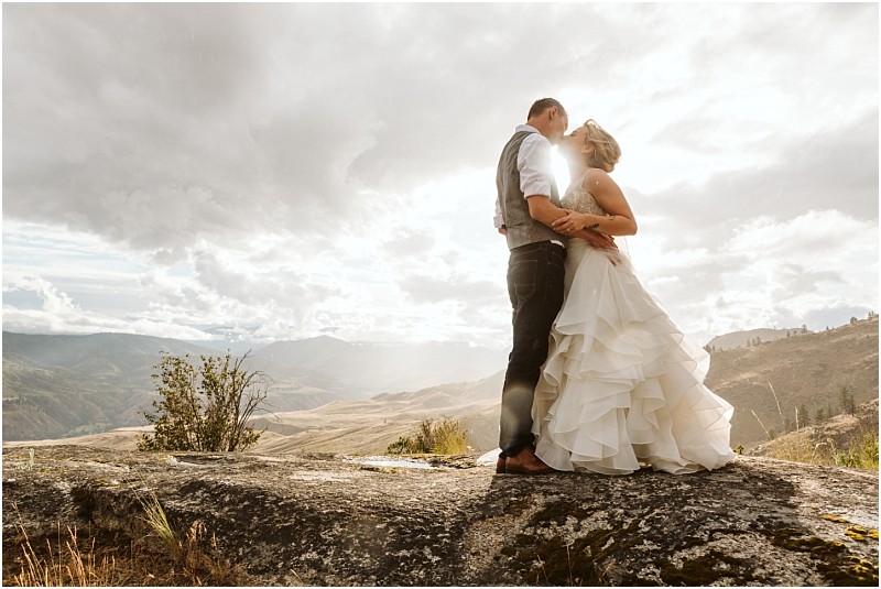 Snohomish & seattle wedding photographer | gsquared weddings photography a bride and groom embrace and kiss on a rocky hilltop during their methow elopement with dramatic clouds and sunlight illuminating rolling mountains and valleys in the background | wedding photo by gsquared weddings