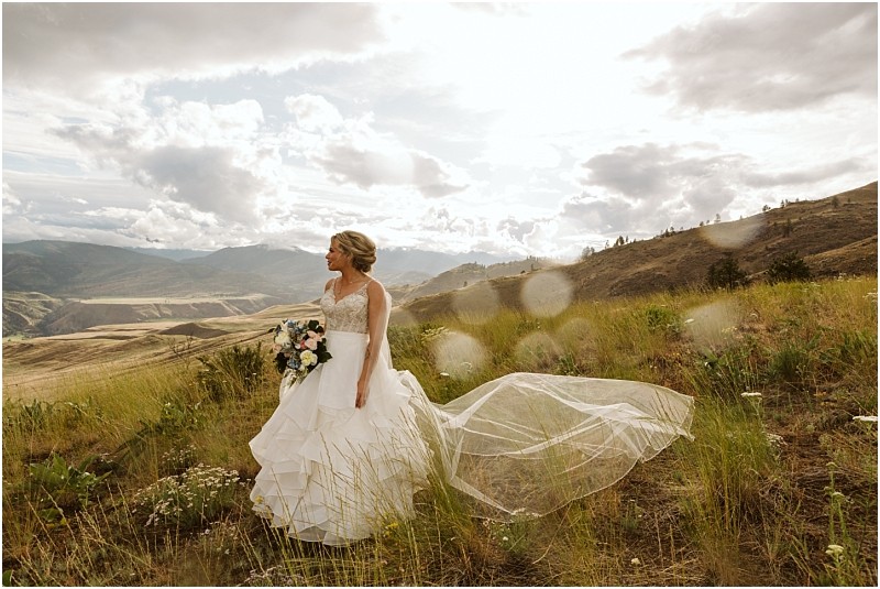 Snohomish & seattle wedding photographer | gsquared weddings photography a bride in a white wedding dress stands in a grassy field on a hillside holding a bouquet and looking to the side during her methow elopement with mountains and dramatic clouds behind her as sunlight shines through creating lens flares | wedding photo by gsquared weddings