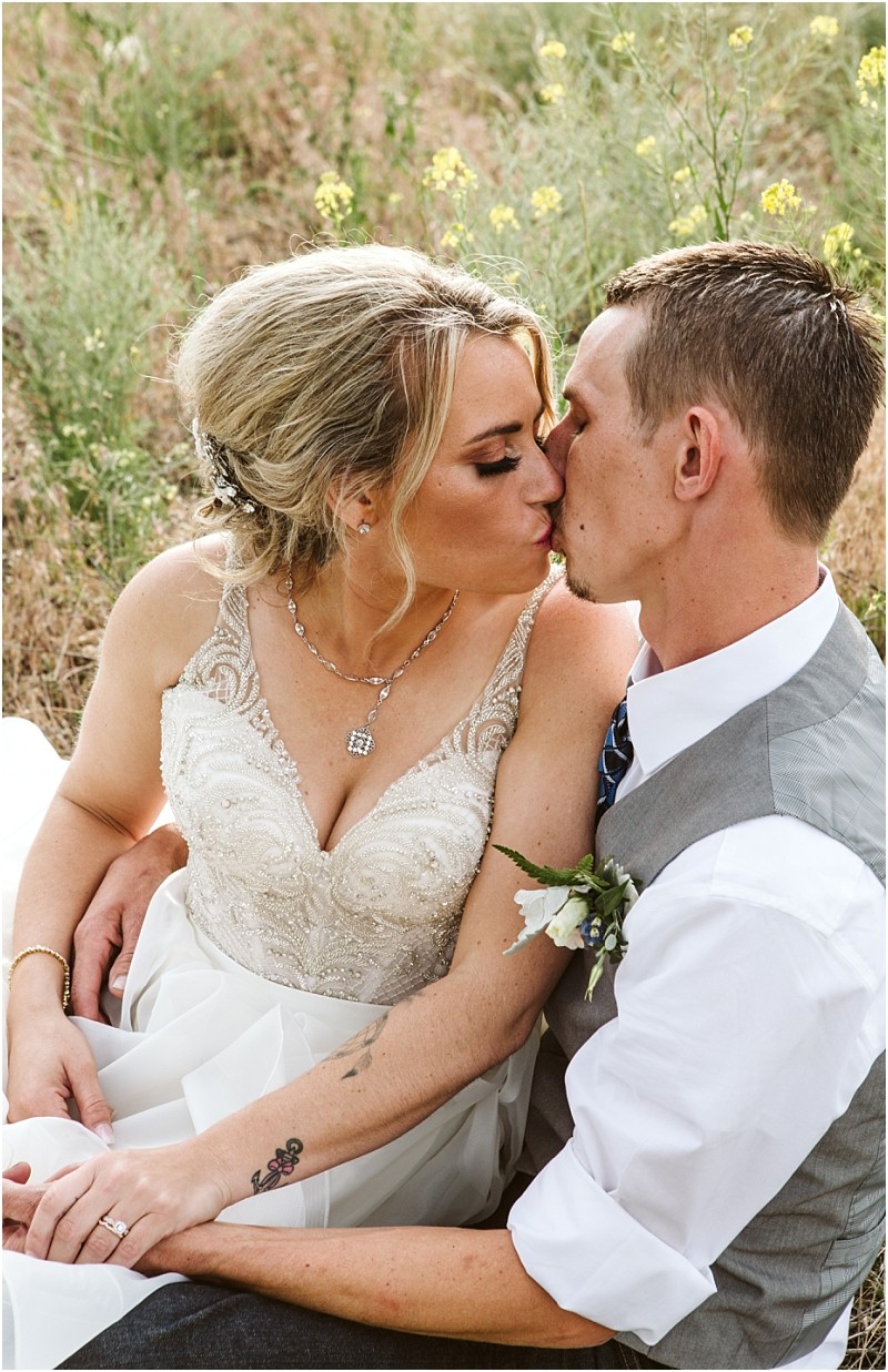 Snohomish & seattle wedding photographer | gsquared weddings photography a bride and groom sit closely together outdoors in a grassy field holding hands and sharing a gentle kiss during their methow elopement The bride wears a beaded dress and necklace while the groom is in a vest with a boutonniere | wedding photo by gsquared weddings