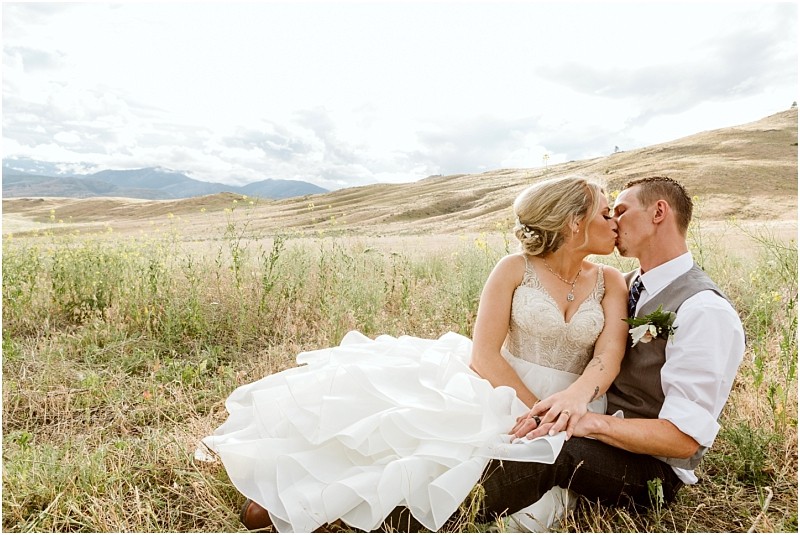 Snohomish & seattle wedding photographer | gsquared weddings photography a bride and groom sit in a grassy field kissing during their methow elopement The bride wears a white dress and the groom is in a vest and tie with rolling hills and a cloudy sky creating a serene outdoor wedding scene | wedding photo by gsquared weddings
