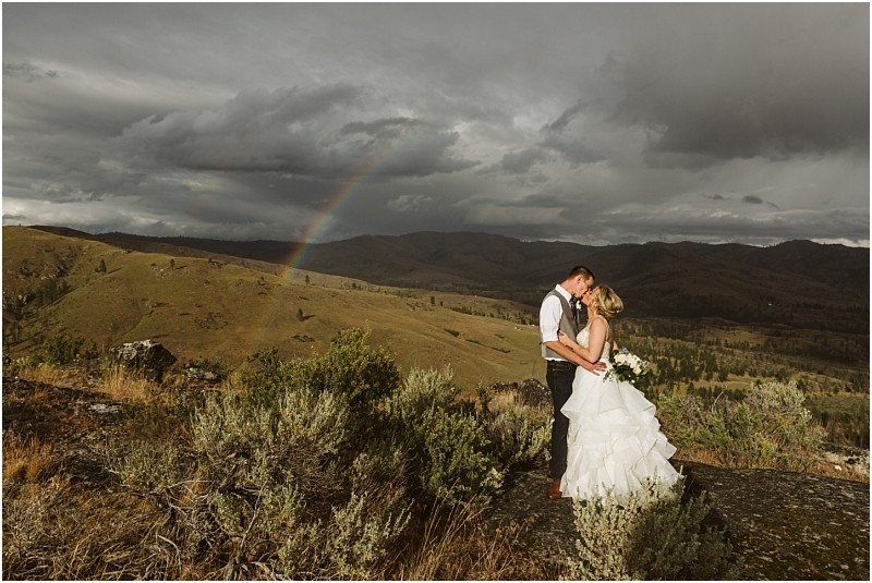 Snohomish & seattle wedding photographer | gsquared weddings photography a bride and groom embrace on a grassy hillside during their methow elopement beneath a dramatic cloudy sky and a rainbow arching over distant mountains The bride wears white and holds a bouquet the groom wears a vest and tie | wedding photo by gsquared weddings