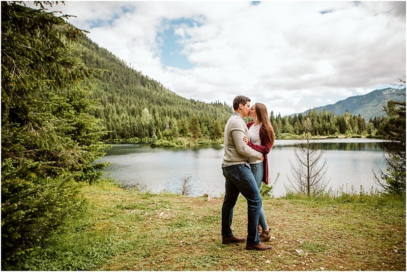 Snohomish & seattle wedding photographer | gsquared weddings photography a couple stands close together about to kiss by gold creek pond a serene lake surrounded by pine trees and mountains under a partly cloudy skycapturing the perfect engagement moment | wedding photo by gsquared weddings
