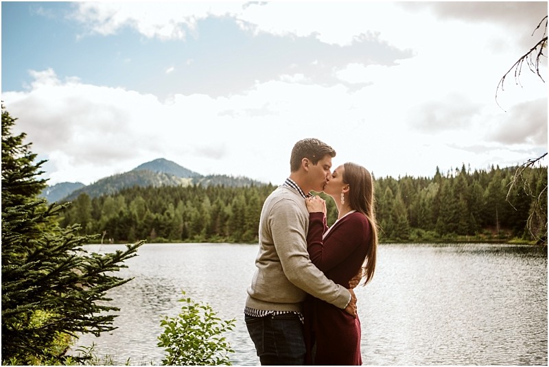 Snohomish & seattle wedding photographer | gsquared weddings photography a couple stands by gold creek pond embracing and sharing a kiss with forested mountains and a cloudy sky in the backgroundcapturing a romantic engagement moment | wedding photo by gsquared weddings