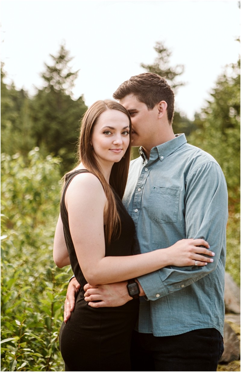 Snohomish & seattle wedding photographer | gsquared weddings photography a couple stands outdoors among greenery at gold creek pond the woman faces the camera with a slight smile while the man wearing a blue shirt hugs her and whispers in her ear Trees are visible in the background capturing their engagement moment | wedding photo by gsquared weddings