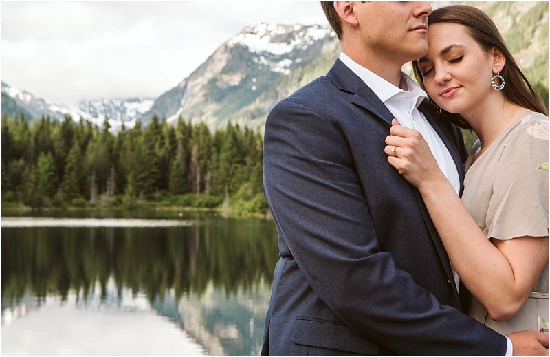 Snohomish & seattle wedding photographer | gsquared weddings photography a couple embraces by calm gold creek pond surrounded by pine trees and snow capped mountains The woman rests her head on the mans chest eyes closed appearing peaceful and content during their engagement moment | wedding photo by gsquared weddings