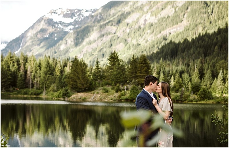 Snohomish & seattle wedding photographer | gsquared weddings photography a couple embraces and kisses by a calm mountain lake at their gold creek pond engagement surrounded by pine trees and tall mountains with patches of snow in the background | wedding photo by gsquared weddings