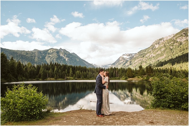 Snohomish & Seattle Wedding Photographer | GSquared Weddings Photography A couple stands embracing on the shore of Gold Creek Pond, surrounded by pine trees and mountains under a partly cloudy sky, their reflection mirrored in the calm lake—capturing a perfect engagement moment.                                   | wedding photo by GSquared Weddings