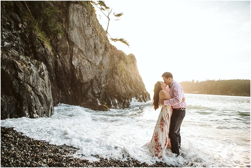 Snohomish & seattle wedding photographer | gsquared weddings photography a couple stands in the shallow waves at the edge of a rocky beach embracing and kissing as the sun sets behind cliffs and trees during their deception pass engagement session in june 2020 | wedding photo by gsquared weddings