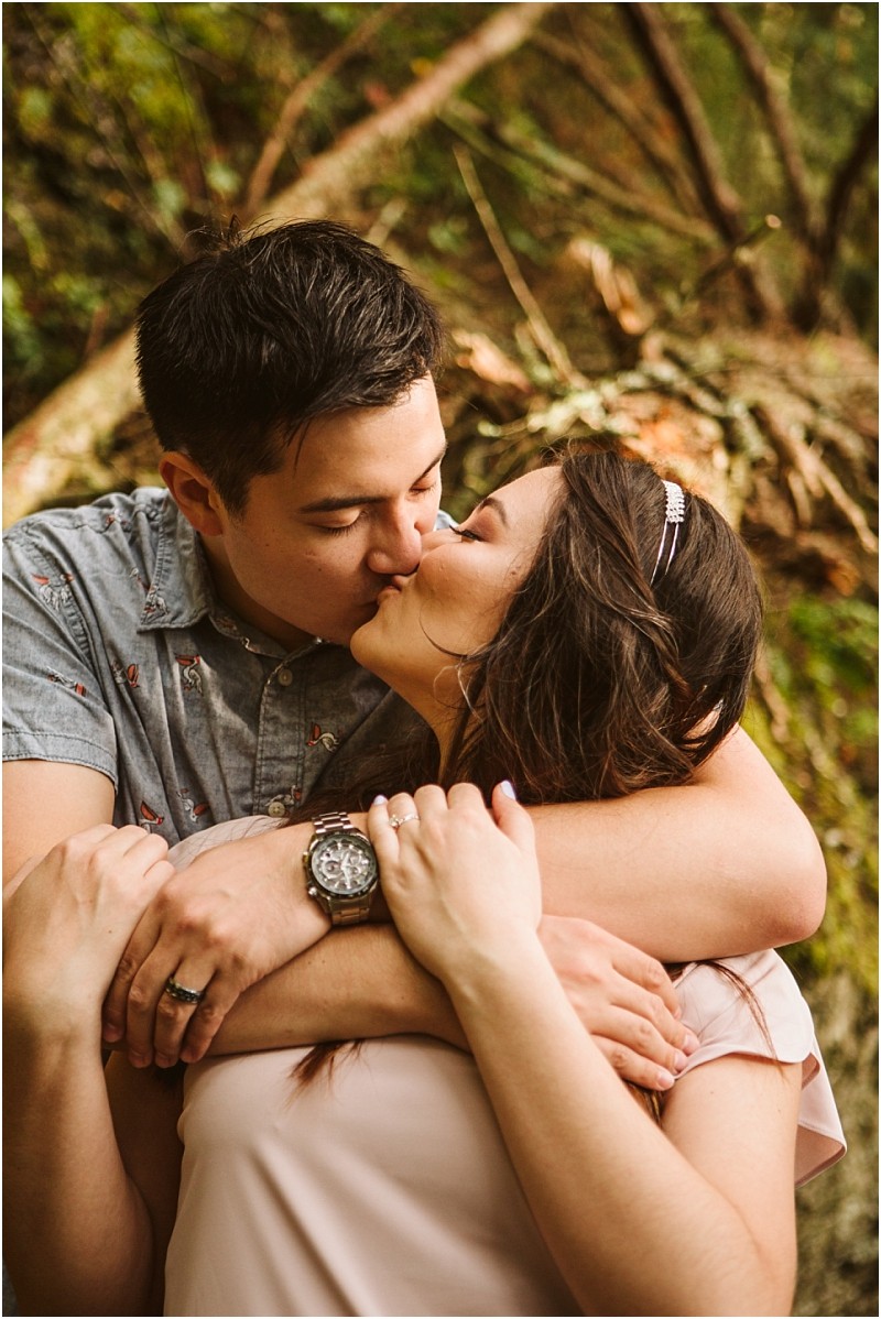 Snohomish & seattle wedding photographer | gsquared weddings photography a couple embraces and kisses outdoors during their deception pass engagement session in june 2020 with the man wrapping his arms around the woman from behind Both appear happy and affectionate surrounded by greenery and fallen branches | wedding photo by gsquared weddings