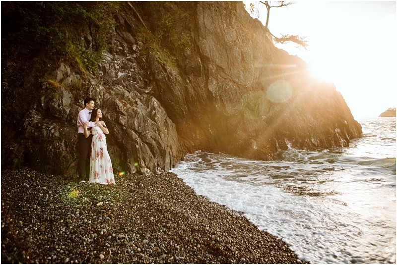 Snohomish & seattle wedding photographer | gsquared weddings photography a couple stands on a rocky beach at sunset during their deception pass engagement session with waves lapping at the shore and sunlight streaming over a cliff behind them The woman wears a floral dress as they gaze together toward the horizon | wedding photo by gsquared weddings