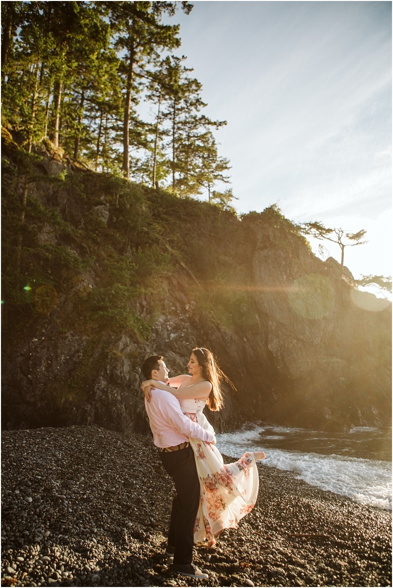 Snohomish & seattle wedding photographer | gsquared weddings photography a man lifts a woman in a floral dress as they smile at each other on a rocky beach during their deception pass engagement session in june 2020 with sunlight streaming through tall trees and cliffs in the background | wedding photo by gsquared weddings