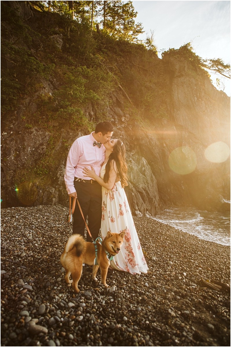 Snohomish & seattle wedding photographer | gsquared weddings photography a couple kisses on a rocky beach at sunset during their deception pass engagement session in june 2020 holding a dog on a leash Sunlight filters through trees as she wears a white floral dress and he sports a pink shirt and dark pants | wedding photo by gsquared weddings