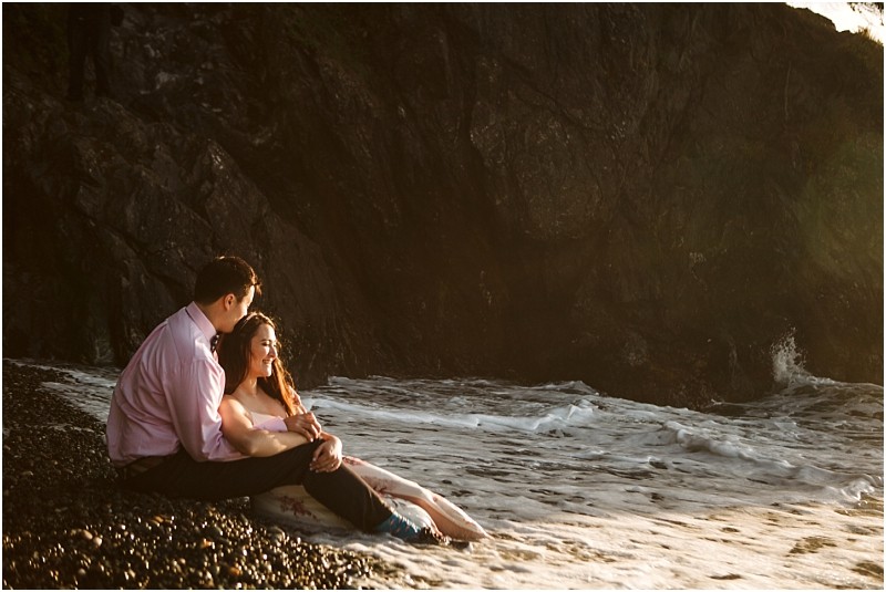 Snohomish & seattle wedding photographer | gsquared weddings photography a couple sits close together on a rocky beach at the waters edge during their deception pass engagement session in june 2020 with gentle waves and sunlight illuminating their faces as rugged cliffs rise behind them | wedding photo by gsquared weddings