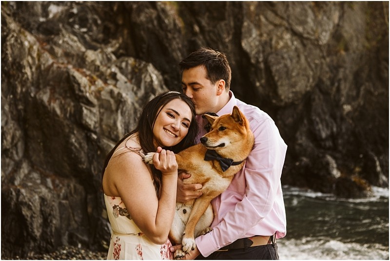 Snohomish & seattle wedding photographer | gsquared weddings photography a smiling woman in a floral dress hugs a man in a pink shirt who kisses her head while holding their shiba inu in a bow tie They stand by the water and rocky cliffs during their deception pass engagement session june 2020 | wedding photo by gsquared weddings