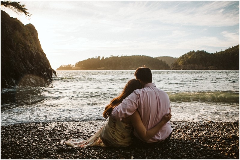 Snohomish & seattle wedding photographer | gsquared weddings photography a couple sits closely together on a rocky beach at deception pass during their june 2020 engagement session facing the ocean as the sun sets behind distant forested hills creating a peaceful and intimate atmosphere | wedding photo by gsquared weddings