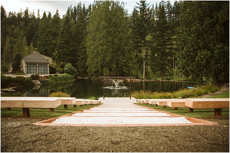 Snohomish & seattle wedding photographer | gsquared weddings photography a wide stone path with wooden benches on both sides leads down to a small pond with a fountain and a graceful gray bridge surrounded by tall trees and a house to the left The scene is peaceful and green | wedding photo by gsquared weddings