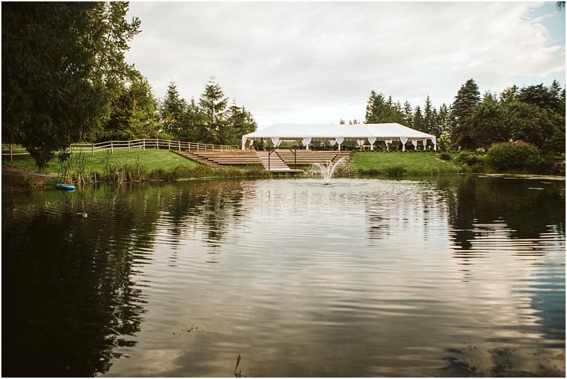 Snohomish & seattle wedding photographer | gsquared weddings photography a white event tent is set up beside a pond with a small fountain surrounded by trees and greenery under a partly cloudy sky A gray bridge arches gracefully near the wooden fence along the grassy hill by the tent | wedding photo by gsquared weddings