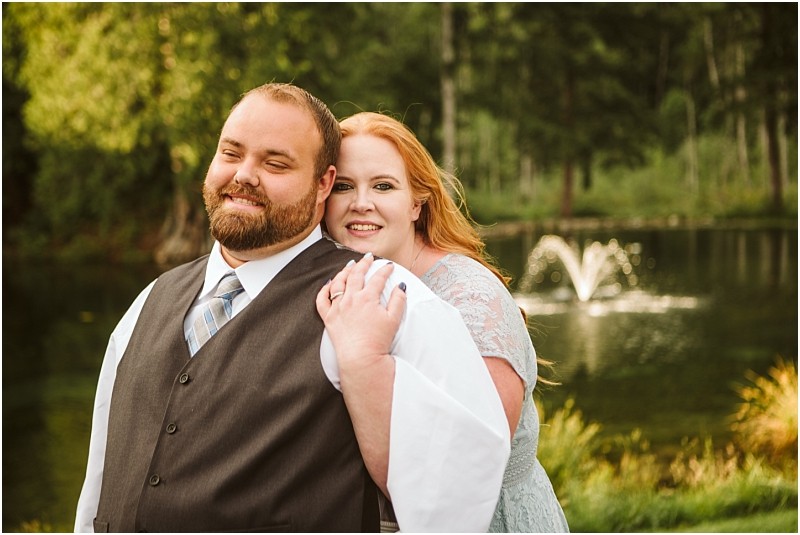 Snohomish & seattle wedding photographer | gsquared weddings photography a smiling couple poses outdoors by a pond with a fountain for their gray bridge engagement The man wears a vest and tie while the woman in a light blue dress embraces him as trees and greenery fill the background | wedding photo by gsquared weddings