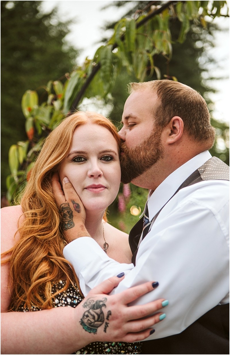 Snohomish & seattle wedding photographer | gsquared weddings photography a man with a beard kisses a red haired woman on the forehead near a gray bridge gently holding her face Dressed formally outdoors with greenery behind them they share an intimate moment tattoos are visible on their hands | wedding photo by gsquared weddings