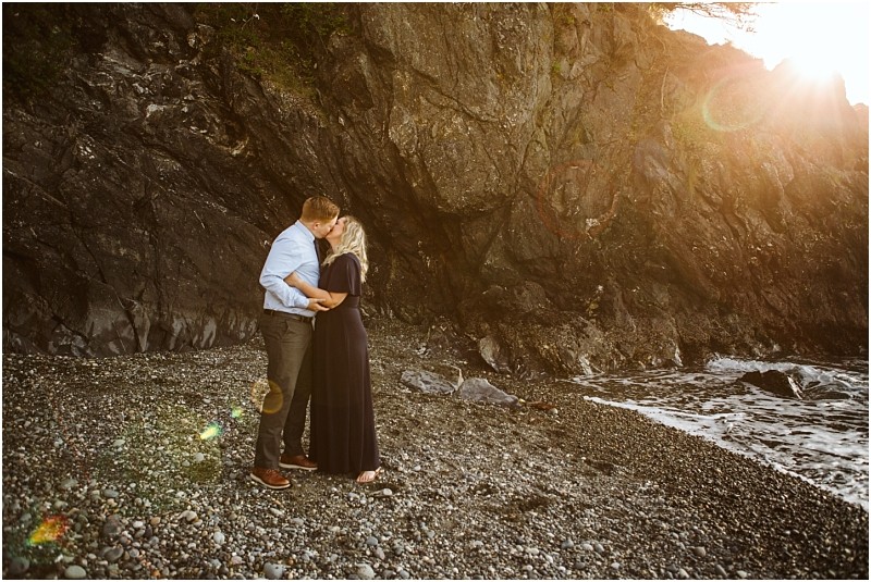 Snohomish & seattle wedding photographer | gsquared weddings photography a couple stands on a rocky beach at sunset near deception pass sharing a kiss by the waters edge Sunlight casts a warm glow on them and the cliff behind capturing the magic of their whidbey island engagement | wedding photo by gsquared weddings