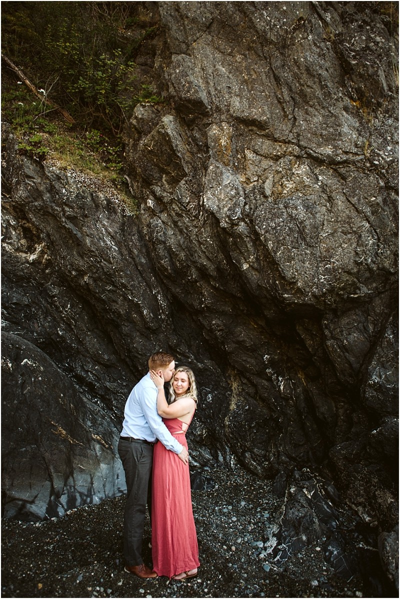 Snohomish & seattle wedding photographer | gsquared weddings photography a couple embraces at the base of a large rocky cliff at deception pass whidbey island The woman in a long pink dress looks at the camera while the man kisses her forehead surrounded by small pebbles | wedding photo by gsquared weddings