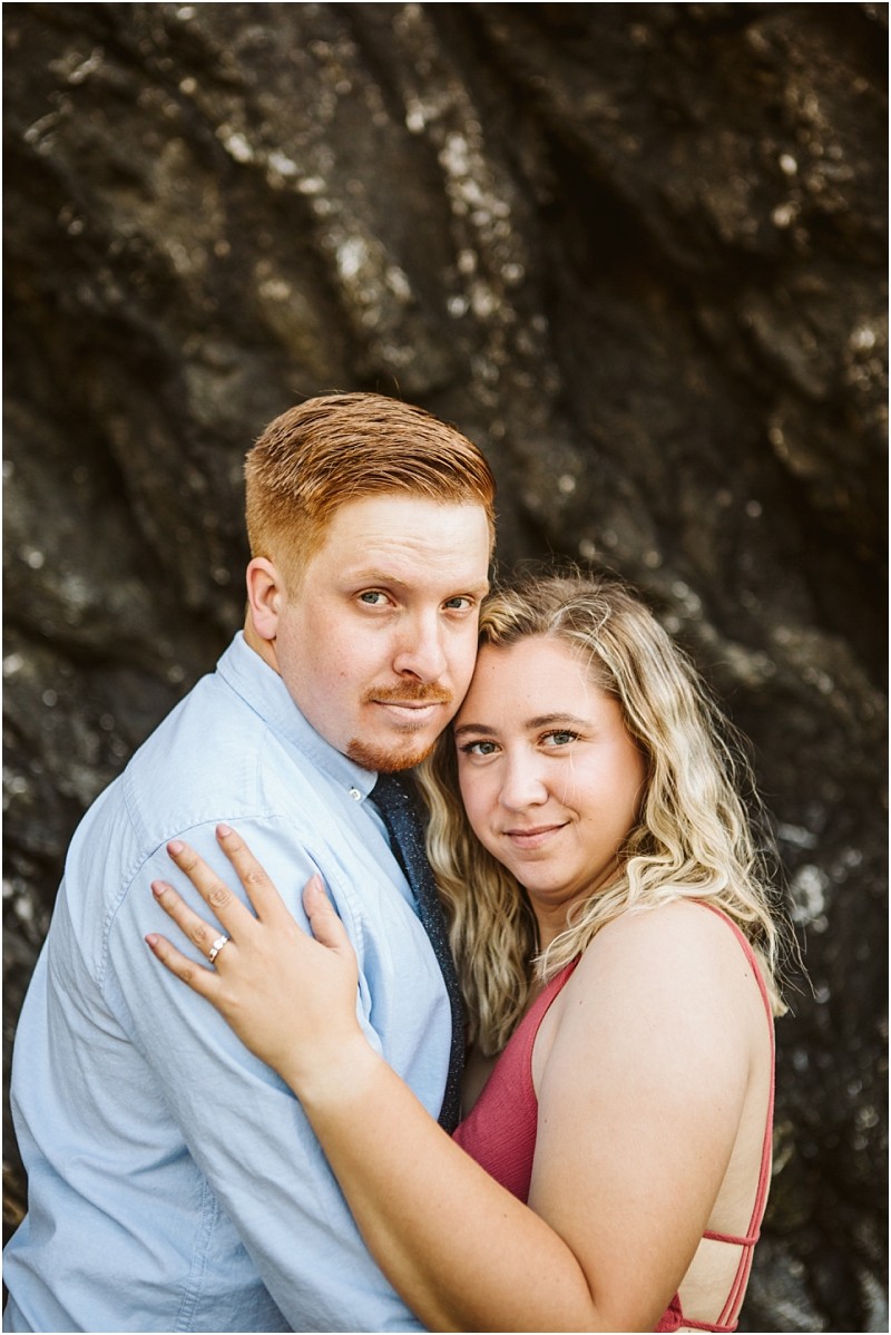 Snohomish & seattle wedding photographer | gsquared weddings photography a couple poses closely together in front of a dark rocky background at deception pass whidbey island The woman with wavy blonde hair wears a sleeveless pink top proudly showing her engagement ring the man wears a light blue shirt and a trimmed beard | wedding photo by gsquared weddings