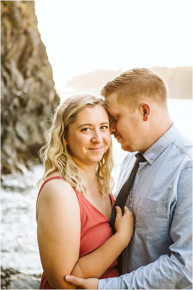 Snohomish & seattle wedding photographer | gsquared weddings photography a couple embraces by the water at deception pass whidbey island the woman in a pink dress smiles at the camera while the man in a light blue shirt and black tie leans his forehead affectionately against her temple with rocky cliffs and water behind them | wedding photo by gsquared weddings