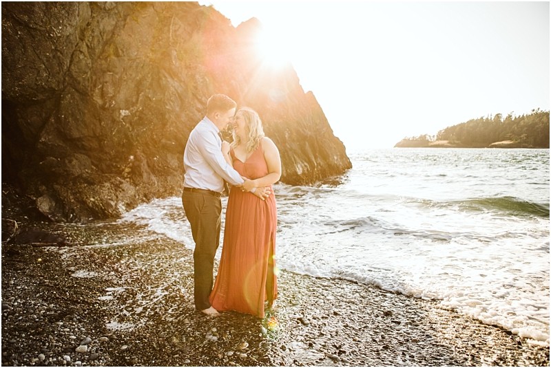 Snohomish & seattle wedding photographer | gsquared weddings photography a couple stands barefoot on a rocky beach at sunset during their deception pass whidbey island engagement embracing and smiling as waves gently touch the shore Sunlight streams over a rocky cliff behind them creating a warm golden glow | wedding photo by gsquared weddings