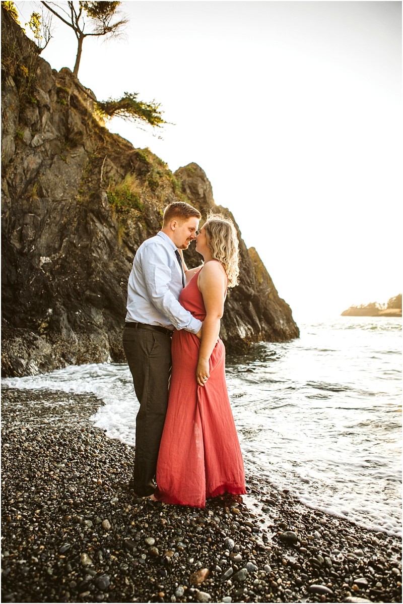 Snohomish & seattle wedding photographer | gsquared weddings photography a couple stands on a rocky beach at sunset embracing during their deception pass whidbey island engagement The woman wears a long coral dress the man a light shirt and dark pants Cliffs water and playful dogs complete the scene | wedding photo by gsquared weddings