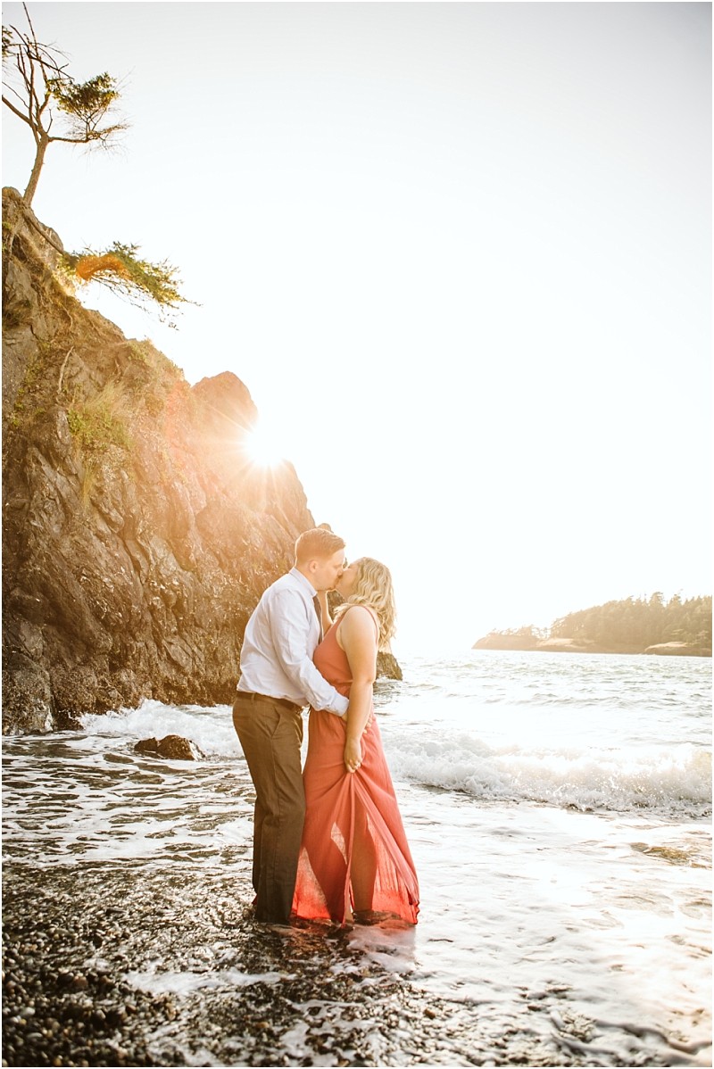 Snohomish & seattle wedding photographer | gsquared weddings photography a couple stands on a rocky beach at sunset at deception pass whidbey island embracing and sharing a kiss as waves wash around their feet The woman in coral and the man in white are framed by sunlight behind a cliff capturing their engagement | wedding photo by gsquared weddings