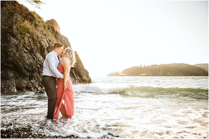 Snohomish & seattle wedding photographer | gsquared weddings photography a couple stands in shallow ocean waves at deception pass embracing and kissing with a rocky cliff and distant islands on whidbey island in the background The woman wears a coral dress the man is in a blue shirtsunlight casting a golden glow | wedding photo by gsquared weddings