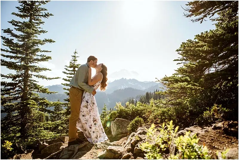 Snohomish & Seattle Wedding Photographer | GSquared Weddings Photography A couple kisses on a rocky mountain trail surrounded by evergreens, sunlight shining and distant peaks in the background. Perfect for a Mount Rainier Tipsoo Engagement Session, she wears a white floral dress, he’s in a green shirt and brown pants. 47.9129° N, 122.0982° W | wedding photo by GSquared Weddings