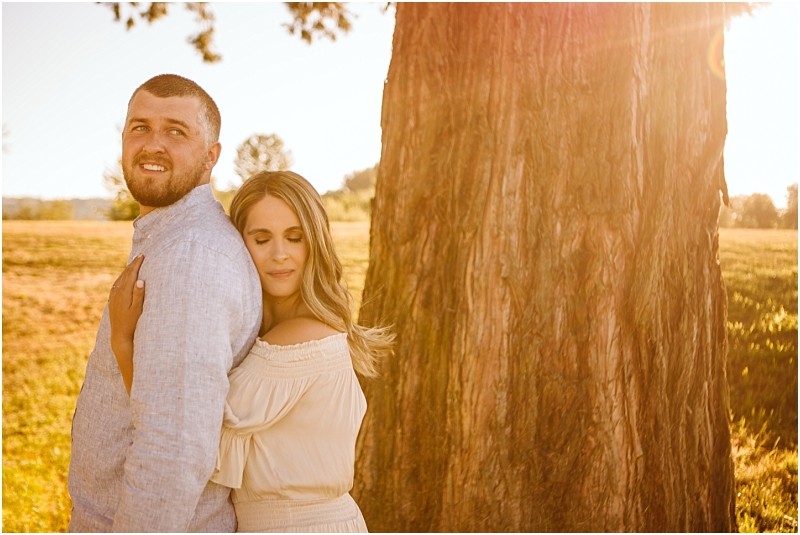 Snohomish & seattle wedding photographer | gsquared weddings photography a couple stands close together by a large tree in a sunlit field at pemberton farm The woman hugs the man from behind resting her head on his backthey look peaceful and relaxed capturing the perfect snohomish engagement moment | wedding photo by gsquared weddings