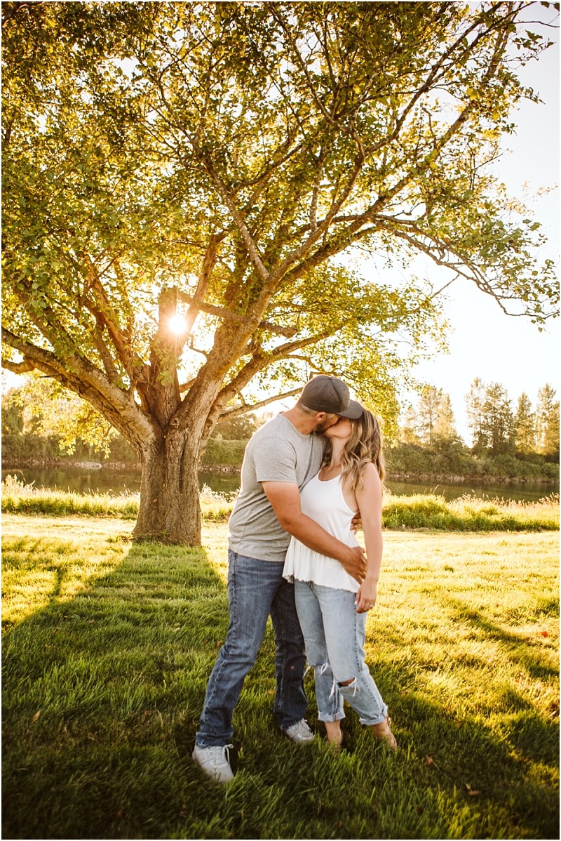 Snohomish & seattle wedding photographer | gsquared weddings photography a couple stands in front of a large tree at sunset kissing and embracing in a sunlit grassy field at pemberton farm They wear casual clothesjeans and t shirtsas sunlight filters through the tree branches behind them in snohomish | wedding photo by gsquared weddings
