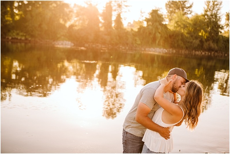 Snohomish & seattle wedding photographer | gsquared weddings photography a couple embraces and kisses by a calm lake at sunset during their pemberton farm engagement with trees and sunlight reflecting on the water in the background | wedding photo by gsquared weddings
