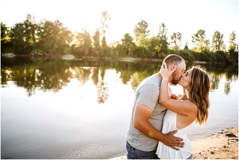 Snohomish & seattle wedding photographer | gsquared weddings photography a couple stands on a sandy riverbank at sunset embracing and kissing The sunlight reflects off the calm water at their pemberton farm engagement in snohomish with trees and greenery in the background Both appear happy and relaxed outdoors | wedding photo by gsquared weddings