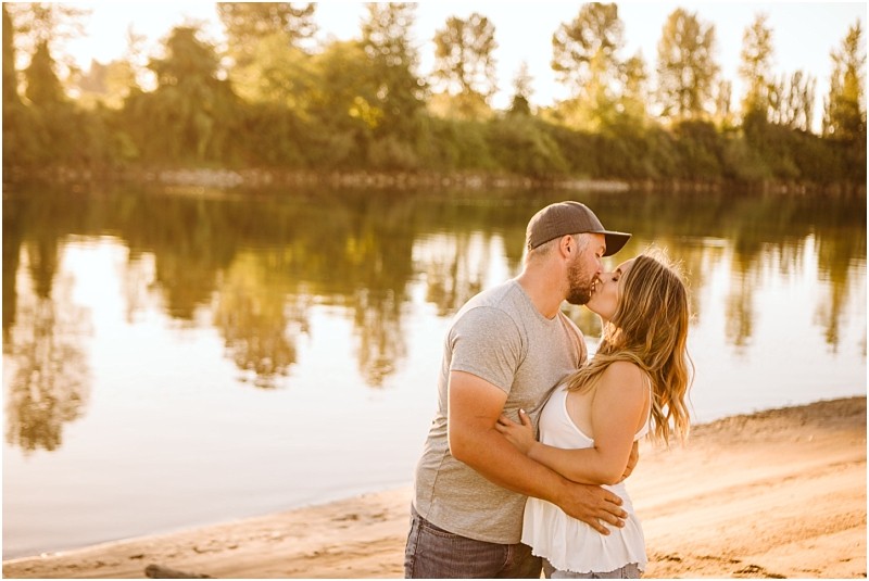 Snohomish & seattle wedding photographer | gsquared weddings photography a couple kisses on a sandy riverbank at sunset during their pemberton farm engagement near snohomish with trees and their reflections visible in the calm water The man wears a cap and grey shirt the woman wears a white sleeveless top | wedding photo by gsquared weddings