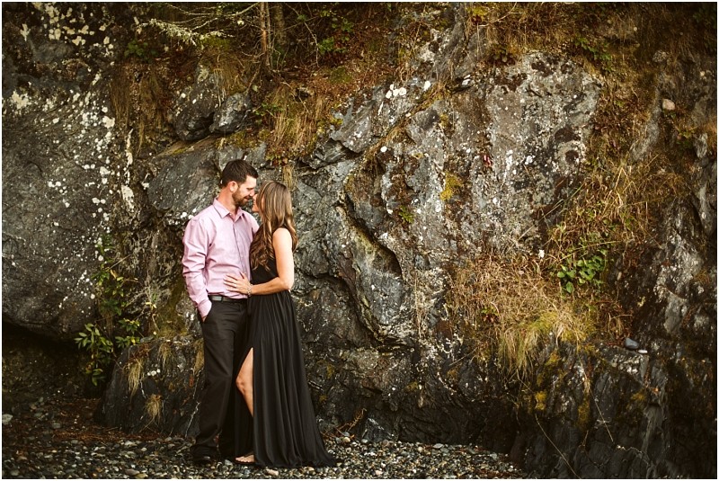 Snohomish & seattle wedding photographer | gsquared weddings photography a couple stands close together on rocky ground at deception pass facing each other in front of a rugged rock wallcapturing the warmth of their august engagement He wears a light pink shirt she stuns in a long black dress with a slit | wedding photo by gsquared weddings