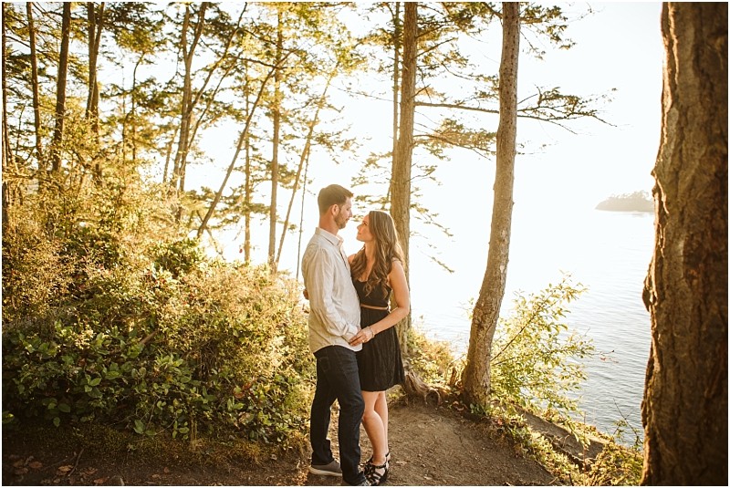 Snohomish & seattle wedding photographer | gsquared weddings photography a couple stands close together in a sunlit forest clearing near deception pass surrounded by tall trees and greenery The woman in a black dress and the man in jeans share an affectionate gaze capturing their august engagement by the water | wedding photo by gsquared weddings