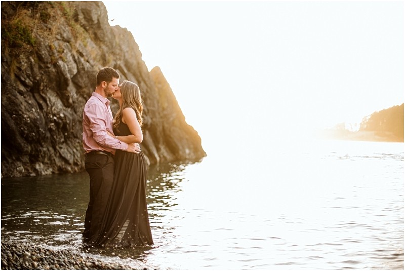 Snohomish & seattle wedding photographer | gsquared weddings photography a couple stands close together at the edge of a rocky shoreline at deception pass partially in the water embracing and looking at each other as the sun setscapturing their august engagement She wears a black dress hes in a pink shirt and dark pants | wedding photo by gsquared weddings
