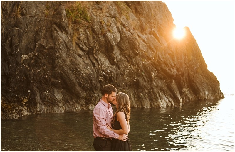 Snohomish & seattle wedding photographer | gsquared weddings photography a couple embraces at the edge of the water near a rocky cliff during their deception pass august engagement with the sun setting behind them and casting a warm golden light over the scene | wedding photo by gsquared weddings