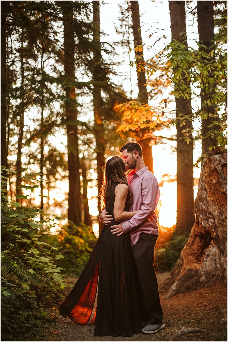 Snohomish & seattle wedding photographer | gsquared weddings photography a couple embraces in a forest at sunset at deception pass with warm sunlight filtering through tall trees The woman wears a long black dress and the man wears a light pink shirtcapturing their magical august engagement amid lush greenery | wedding photo by gsquared weddings