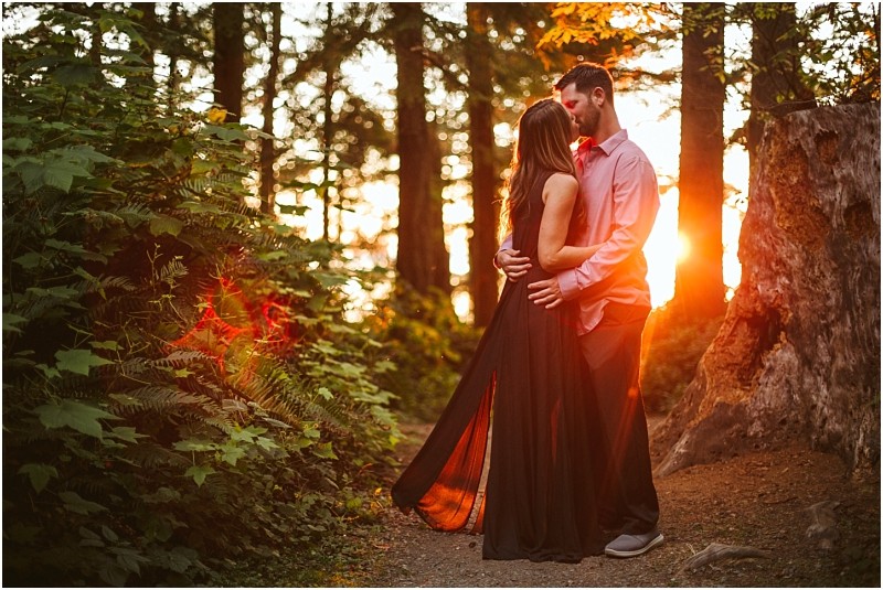 Snohomish & seattle wedding photographer | gsquared weddings photography a couple embraces on a forest path at sunset during their deception pass august engagement Sunlight streams through the trees illuminating the womans long black dress and casting a warm glow around them | wedding photo by gsquared weddings