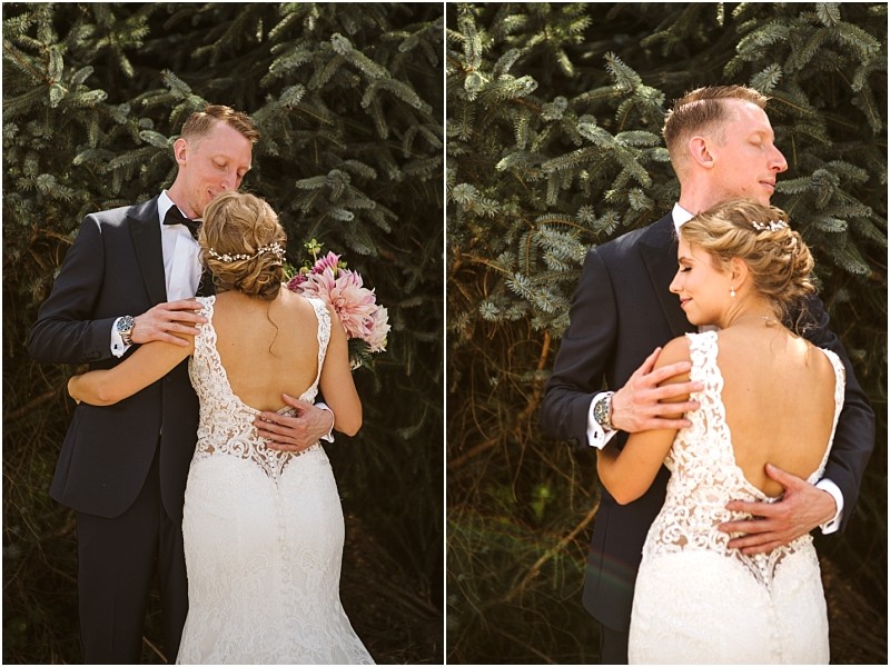 A bride in a white lace gown with an open back hugs her groom in a black tuxedo outdoors both looking emotional and content among the pine trees of snohomish Snohomish wedding photography