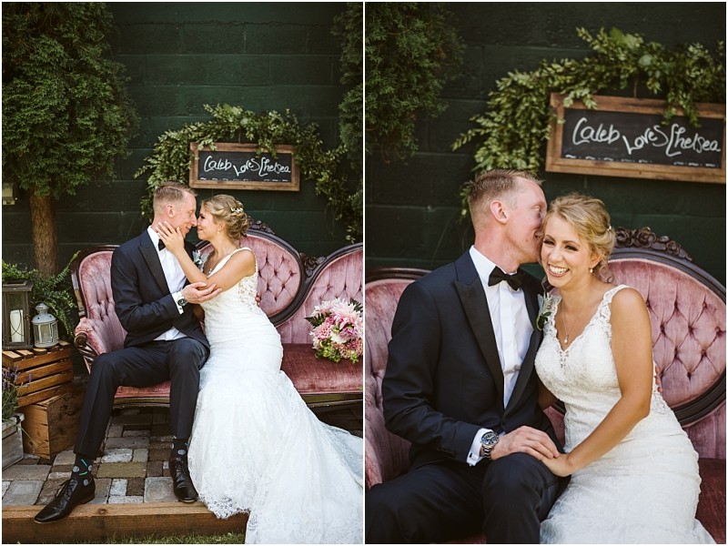 A bride and groom sit on a vintage pink sofa outdoors in snohomish smiling and embracing each other Behind them is a chalkboard sign that reads caleb loves chelsea surrounded by greenery and rustic décor Snohomish wedding photography